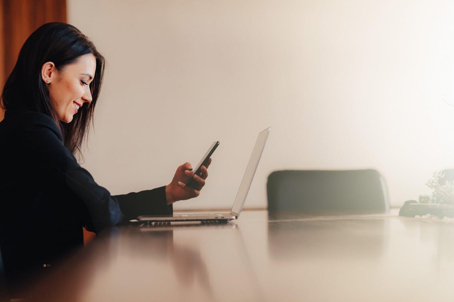 Mulher sorridente usando celular e notebook em ambiente corporativo